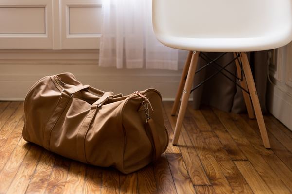 Camel brown duffel bag on stained wooden floor next to an IKEA chair in the corner of a room