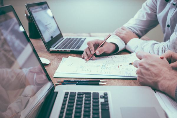 Closeup of two male coworkers looking through some paperwork between two laptops